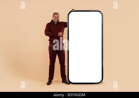 Black man standing beside oversized smartphone mockup and pointing to blank screen Stock Photo