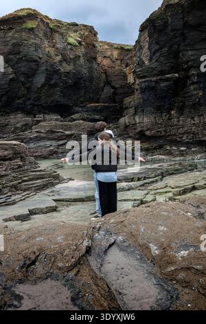 Young couple standing back to back with arms outstretched on rugged beach rocks by dramatic cliffs and cloudy sky, symbolizing partnership, balance an Stock Photo
