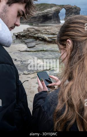Young man and woman on As Catedrais beach are looking at a smartphone, indicating modern travel, navigation, digital communication, and sharing experi Stock Photo