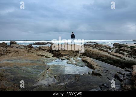 Man standing alone on a rugged rocky coastline under a dramatic cloudy sky, gazing out at the vast ocean waves breaking against the shore, evoking the Stock Photo