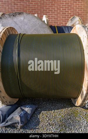 Large wooden cable reel with coiled black and yellow striped wire on gravel near a brick wall; industrial utility equipment outdoors. Stock Photo
