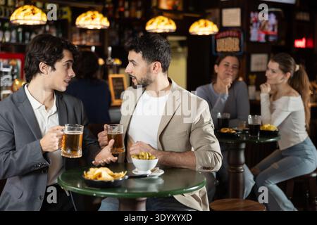 Two handsome positive young guys friends talking and drinking beer at table in beer pub Stock Photo