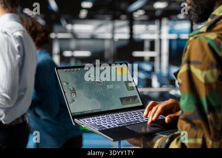 Black army soldier in data center using laptop to overview satellite data via drone control, looking at mission map with command tools. Military expert developing a new safety strategy. Stock Photo