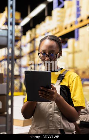Black employee checking delivery records in warehouse logistics system on tablet. Inventory and packaging activities occur within structured shelving setup, ensuring accurate order fulfillment. Stock Photo