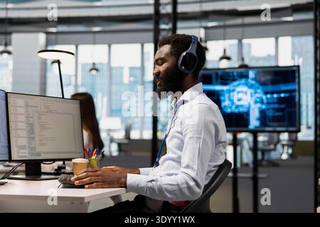 Admin in office using AI automation scripts to reduce repetitive coding tasks while listening music. Employee wearing audio headphones using artificial intelligence technology on PC Stock Photo