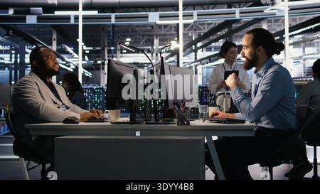 Data center technician discussing with coworker while doing AI software updates. Server room IT specialist and colleague chatting, optimizing infrastructure using artificial intelligence, camera A Stock Photo