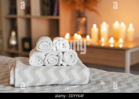Rolled white towels stacked on a bed or massage table, offering a concept of spa, wellness, and relaxation with blurred candles Stock Photo