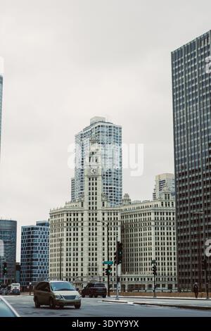 Cars drive on the street near a historic clock tower surrounded by tall buildings in Chicago on a cloudy day. Stock Photo