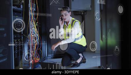 Kneeling IT tech inspecting patch cables, talking on phone in server room, wearing vest with HUD Stock Photo