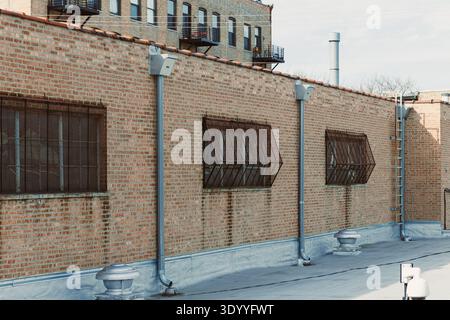 Brick wall has metal grates on windows and ventilation pipes on the rooftop with clear sky in the background Stock Photo