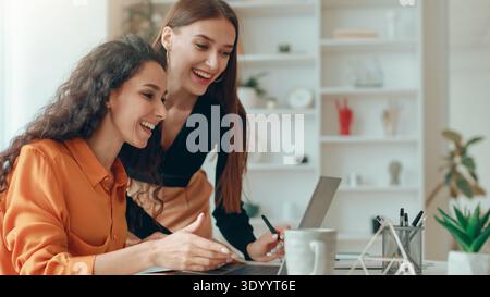 Two Women Collaborating at a Workspace While Smiling and Discussing a Project Together Stock Photo
