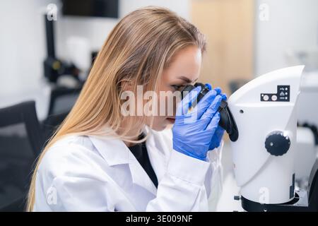 Technician examining samples through high resolution microscope in clinical laboratory. Clean workspace with modern instruments and digital displays visible in the background. Stock Photo