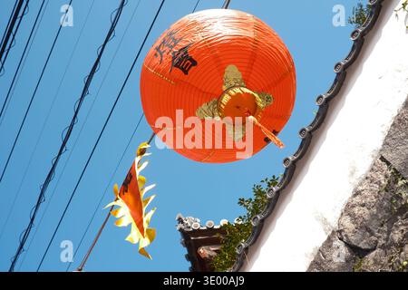 Low angle view of a red Chinese lantern against a clear blue sky and traditional roof tiles, Nagasaki Stock Photo