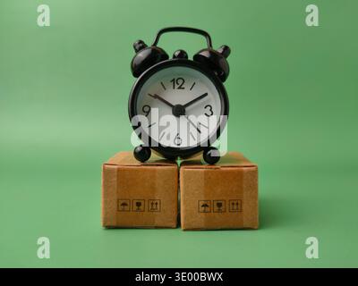 A black vintage-style alarm clock resting on top of two small cardboard parcels against a solid green background. Stock Photo