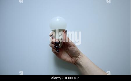A woman's hand with dark nail polish holding a white LED light bulb against a simple light gray background. Concept of energy saving, eco-friendliness, innovation, electricity, and bright ideas. Stock Photo