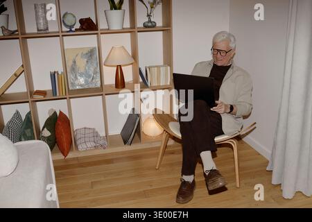 Senior Caucasian Man Sitting in Chair Indoors Using Laptop and Smiling Relaxedly Stock Photo