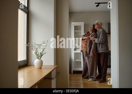 Senior Caucasian Couple Standing Together Smiling and Looking out Window Stock Photo