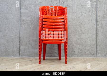 Vertical stack of glossy orange plastic stacking chairs positioned against textured gray concrete wall on light wooden floor indoor, selective focus Stock Photo