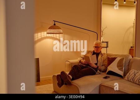 Senior Caucasian Man Sitting on Sofa Reading Book in Cozy Living Room Stock Photo