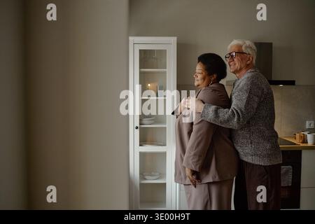 Senior Caucasian Man Embracing Senior Black Woman Standing in Kitchen Stock Photo