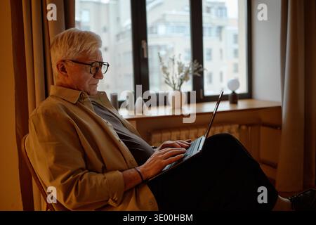 Senior Caucasian Man Using Laptop While Sitting Indoors near Window Stock Photo