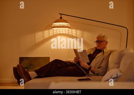 Senior Caucasian Man Relaxing on Sofa Reading Book in Cozy Living Room Stock Photo