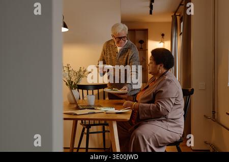 Senior Caucasian Man Serving Food to Senior Black Woman Working on Laptop Stock Photo