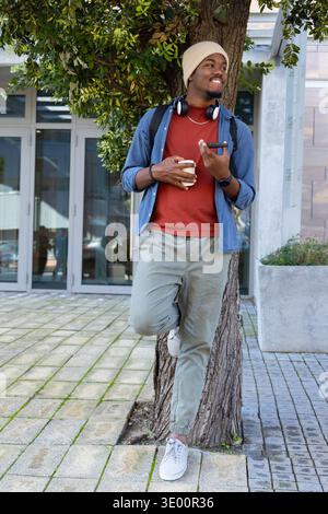 African American man leaning against tree in plaza holding coffee and smartphone with headphones Stock Photo