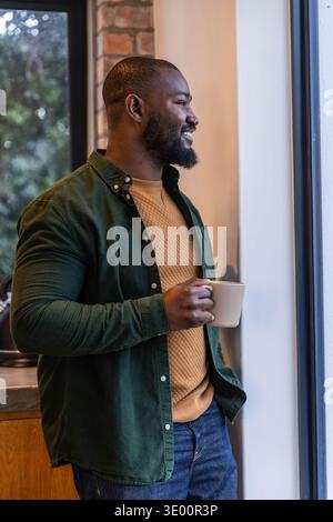 African American man standing in kitchen by big window holding white mug, wearing green shirt Stock Photo
