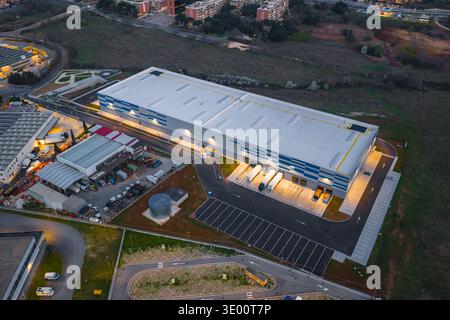 Aerial view of a sprawling warehouse complex gleaming under the evening sky, its loading docks bustling with activity against a backdrop of urban deve Stock Photo