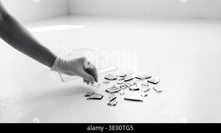 Black and white close-up shot of a protective gloved hand carefully collecting sharp shards of broken glass from a clean. Stock Photo