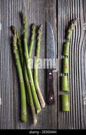 Sliced and whole raw green asparagus spears placed parallel on rustic wood background. Fresh seasonal vegetables in top view Stock Photo