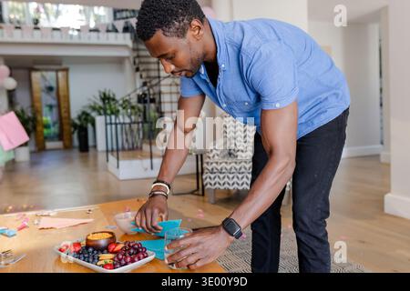 Mid-adult African American man leaning over dining table arranging glass and teal napkins at home. Host, celebration, gathering, fruit, platter, decor Stock Photo