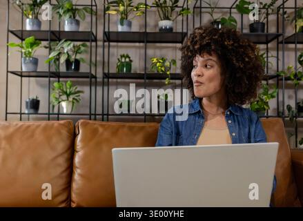African American woman sitting on tan leather sofa working on laptop near plant shelves, copy space. Curly, denim, computer, cozy, greenery, homeoffic Stock Photo
