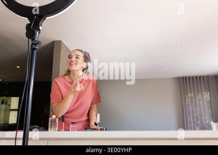 Adult woman wearing coral top and headband at white countertop applying makeup under ring light. Cosmetics, influencer, creator, beauty, studio, home, Stock Photo