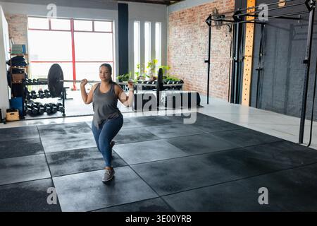 Woman performing forward lunge with loaded barbell on rubber mats in studio building power in tank. Gym, strength, training, weights, industrial, bric Stock Photo