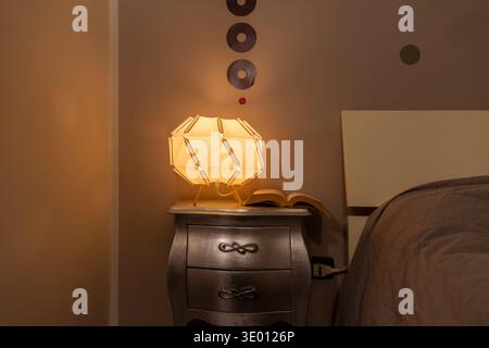Minimal bedroom corner with bedside lamp and books on nightstand, soft warm light creating relaxing atmosphere. Stock Photo