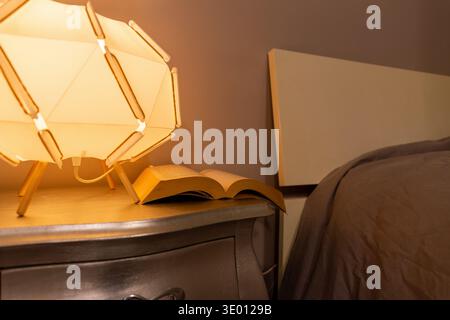 Warm reading lamp illuminating an open book on bedside table in a calm bedroom setting. Stock Photo