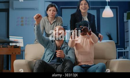 Cheerful coworkers playing video games after work for fun while woman using controller and vr glasses to play game. Workmates enjoying free time after hours with technology at office. Stock Photo