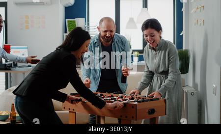 Cheerful woman dancing after winning game at foosball table at office. Coworkers playing football and enjoying fun activity after work. Multi ethnic colleagues having drinks and snacks Stock Photo