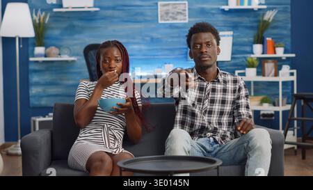 POV of black couple watching television on couch at home relaxing. African american woman eating popcorn and man switching channels with TV remote control while looking at camera Stock Photo