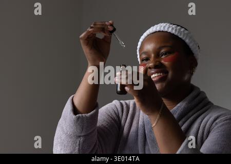 African American woman wearing terry headband gray robe using amber dropper on red patches at home Stock Photo