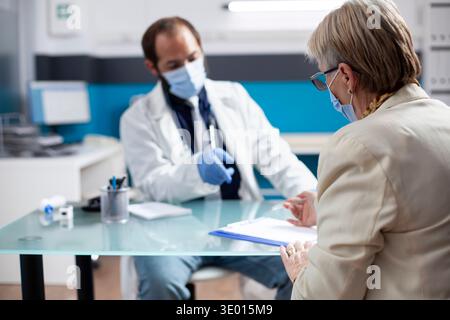 Senior caucasian woman sits in hospital room completing form on clipboard as medic specialist provides guidance. Attentive healthcare worker helps elderly patient navigate medical checkup process. Stock Photo