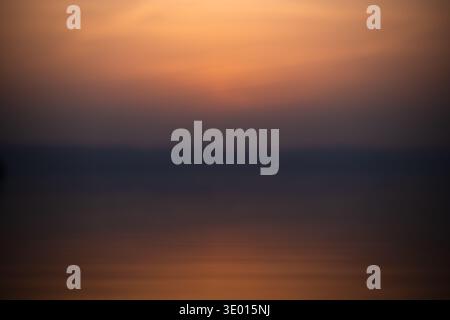 A soft, out of focus image capturing the warm orange and dark blue tones of a sunset reflecting on a calm lake, creating a dreamy and minimal abstract Stock Photo