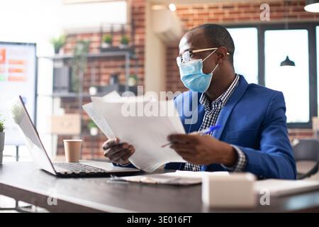 Black businessman holding financial documents and comparing the information on laptop in startup office. Male entrepreneur reviews company research data, using digital device and paperwork. Stock Photo