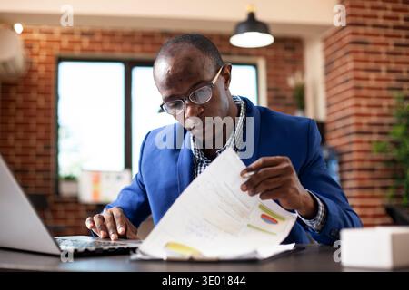 Closeup of black man seated at office desk, deeply focused on analyzing financial documents in brick wall room. African american male manager examining graphs on clipboard with laptop nearby. Stock Photo