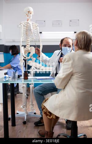 Bearded doctor discusses human anatomy with senior woman, gesturing toward skeleton in clinic office. Retired lady sitting at desk as masked physician provides medical advice using skeleton in clinic. Stock Photo