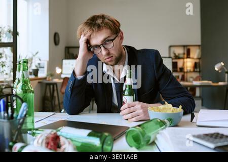 Young Caucasian Man Sitting at Desk Drinking Beer and Looking Stressed Stock Photo