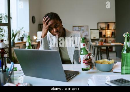 Young Adult Black Woman Sitting at Desk Drinking Beer and Looking Stressed Stock Photo