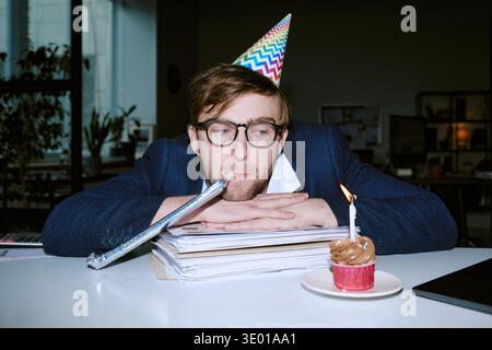 Young Caucasian Man Wearing Party Hat Sitting at Desk Celebrating Birthday Stock Photo